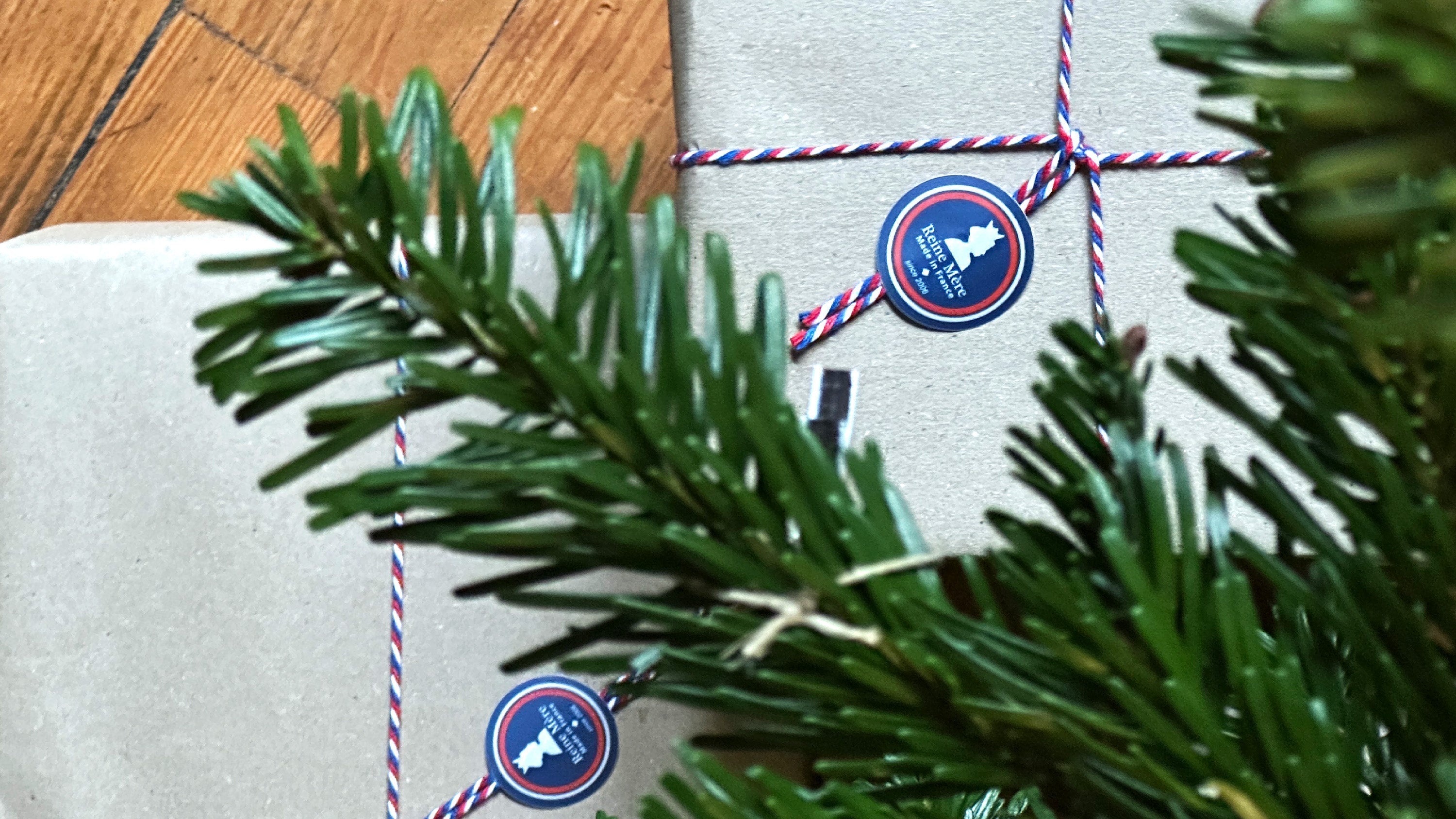 Cadeau emballé en papier kraft avec ficelle tricolore et étiquette Reine Mère, entouré de branches de sapin
